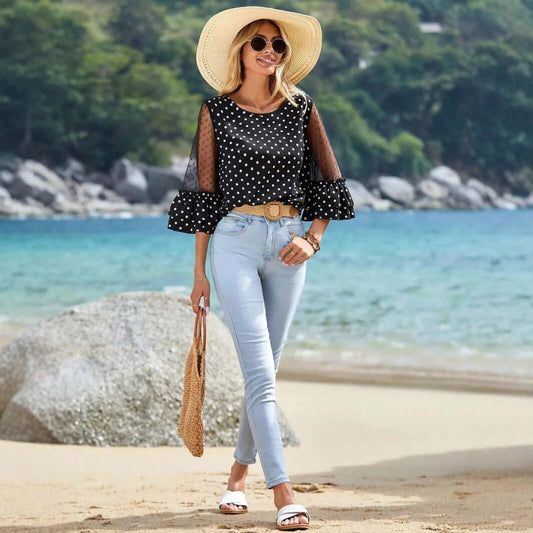 Woman wearing Black Polka Dot Sheer Blouse with 3/4 bell sleeves, paired with light jeans and sun hat, strolling on the beach.