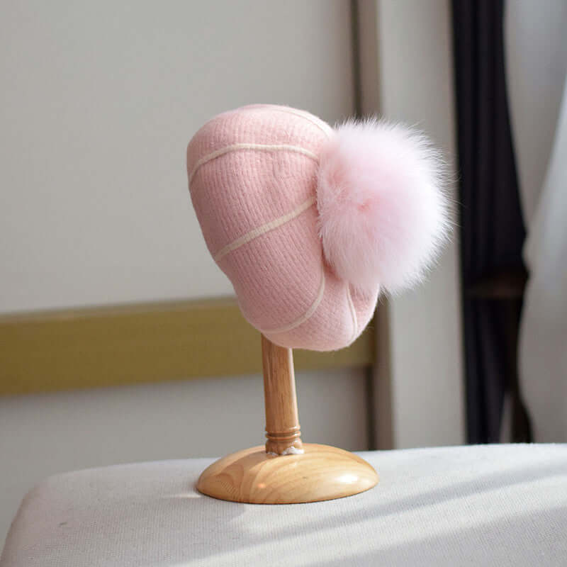Pink beanie with a fluffy fur ball displayed on a wooden stand, perfect for winter style and warmth.