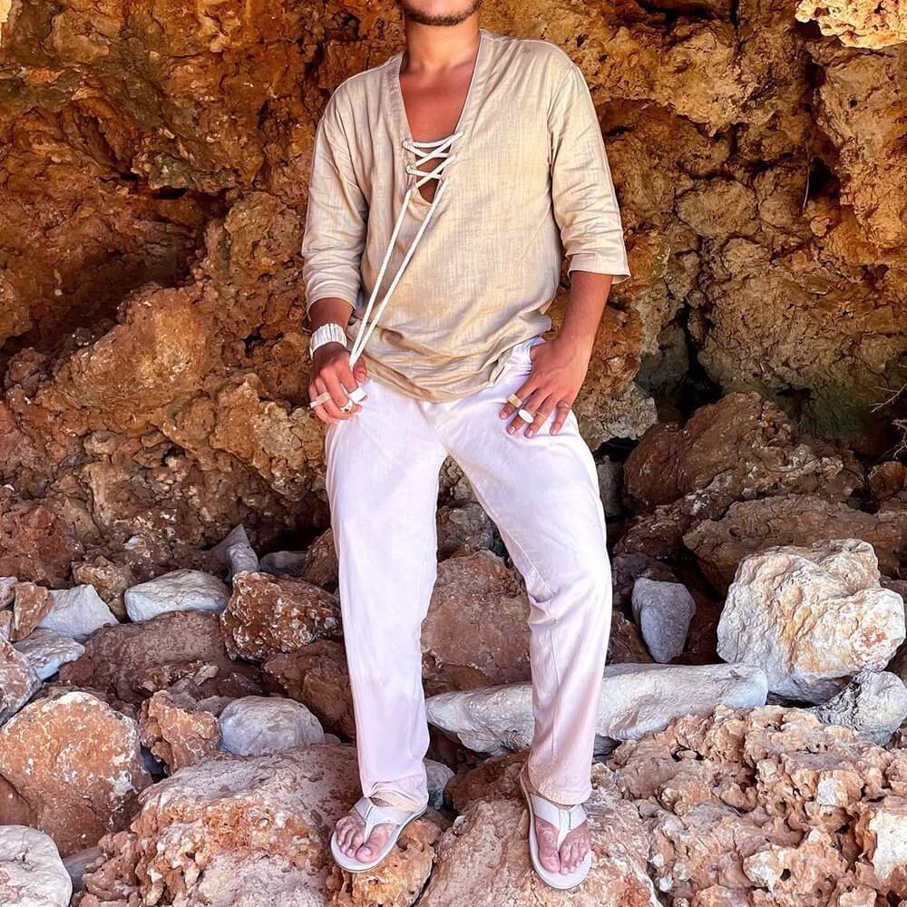 Man wearing a beige lace up T-shirt standing against a rocky background, showcasing casual and stylish outdoor fashion.