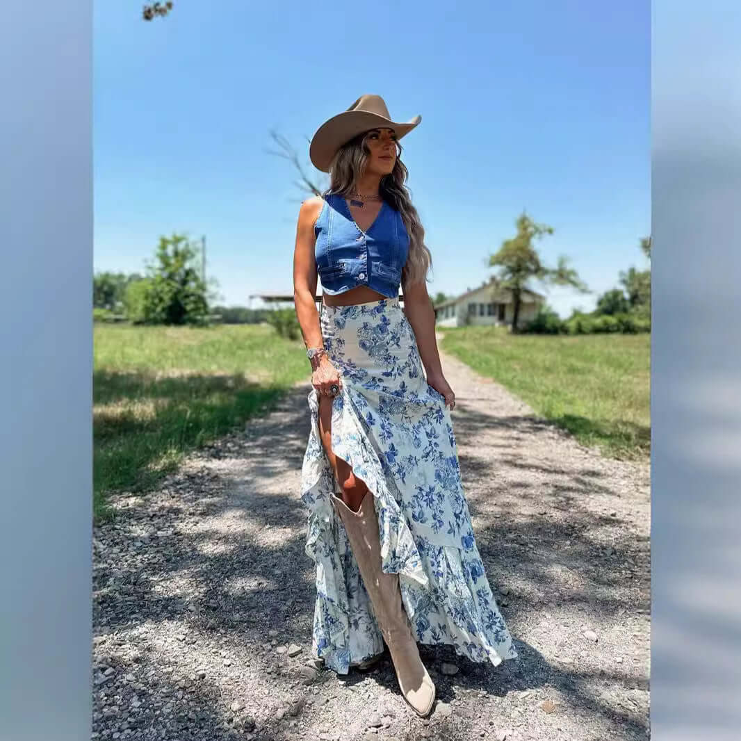 Woman in a floral skirt and denim vest, wearing a hat in a rustic outdoor setting.