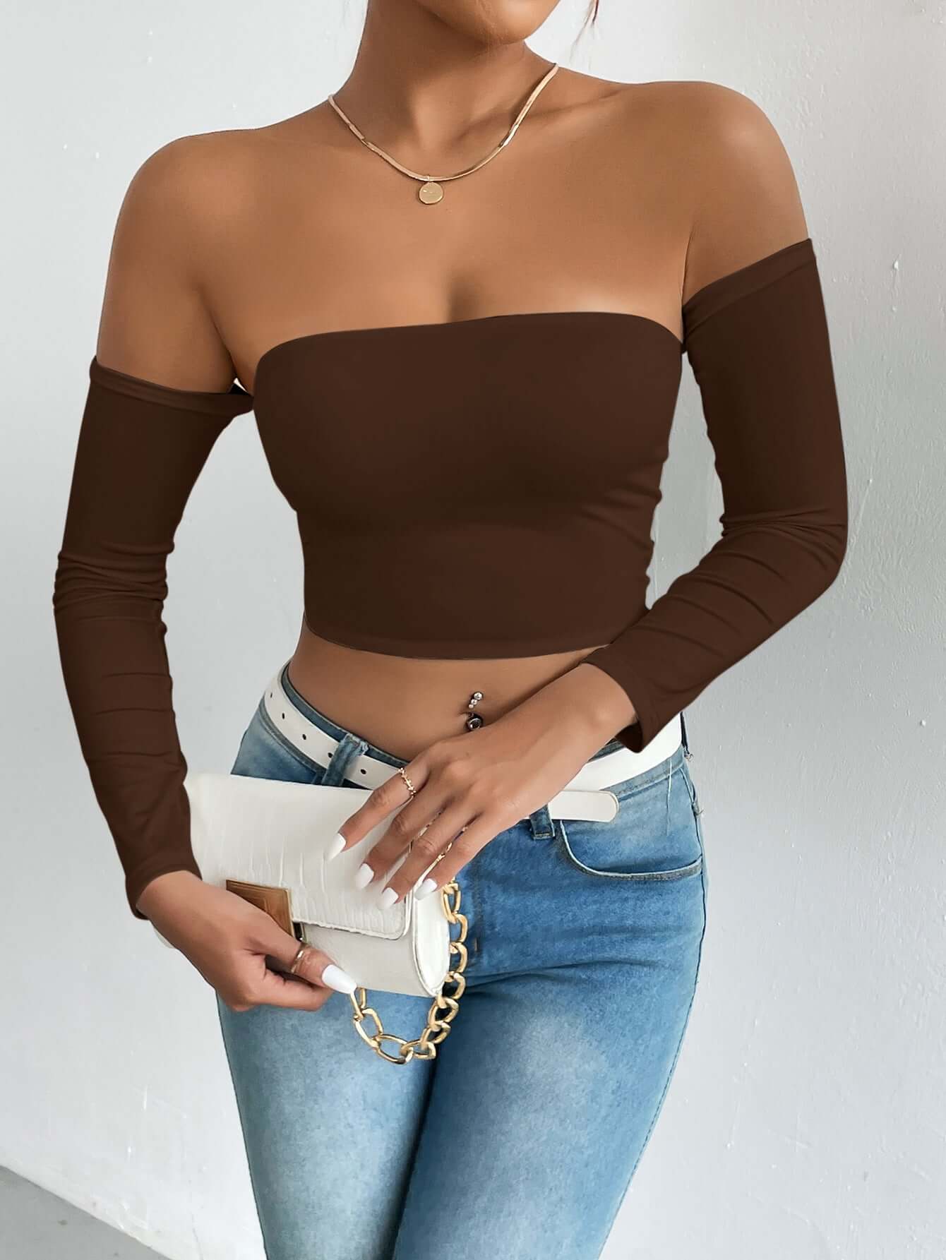 Woman wearing a brown off-shoulder crop top with long sleeves, holding a stylish white clutch bag and wearing blue jeans.