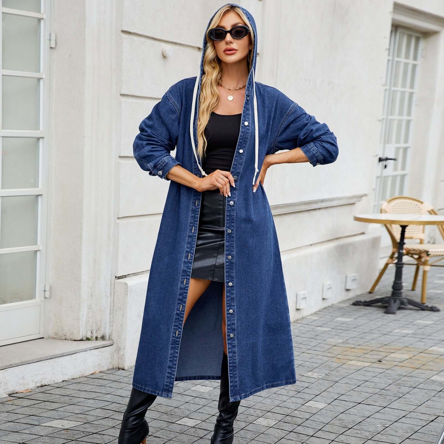 Woman wearing a long blue denim jacket with hood, styled with black outfit underneath, standing outside by a cafe.