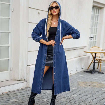 Woman wearing a long blue denim jacket with hood, styled with black outfit underneath, standing outside by a cafe.