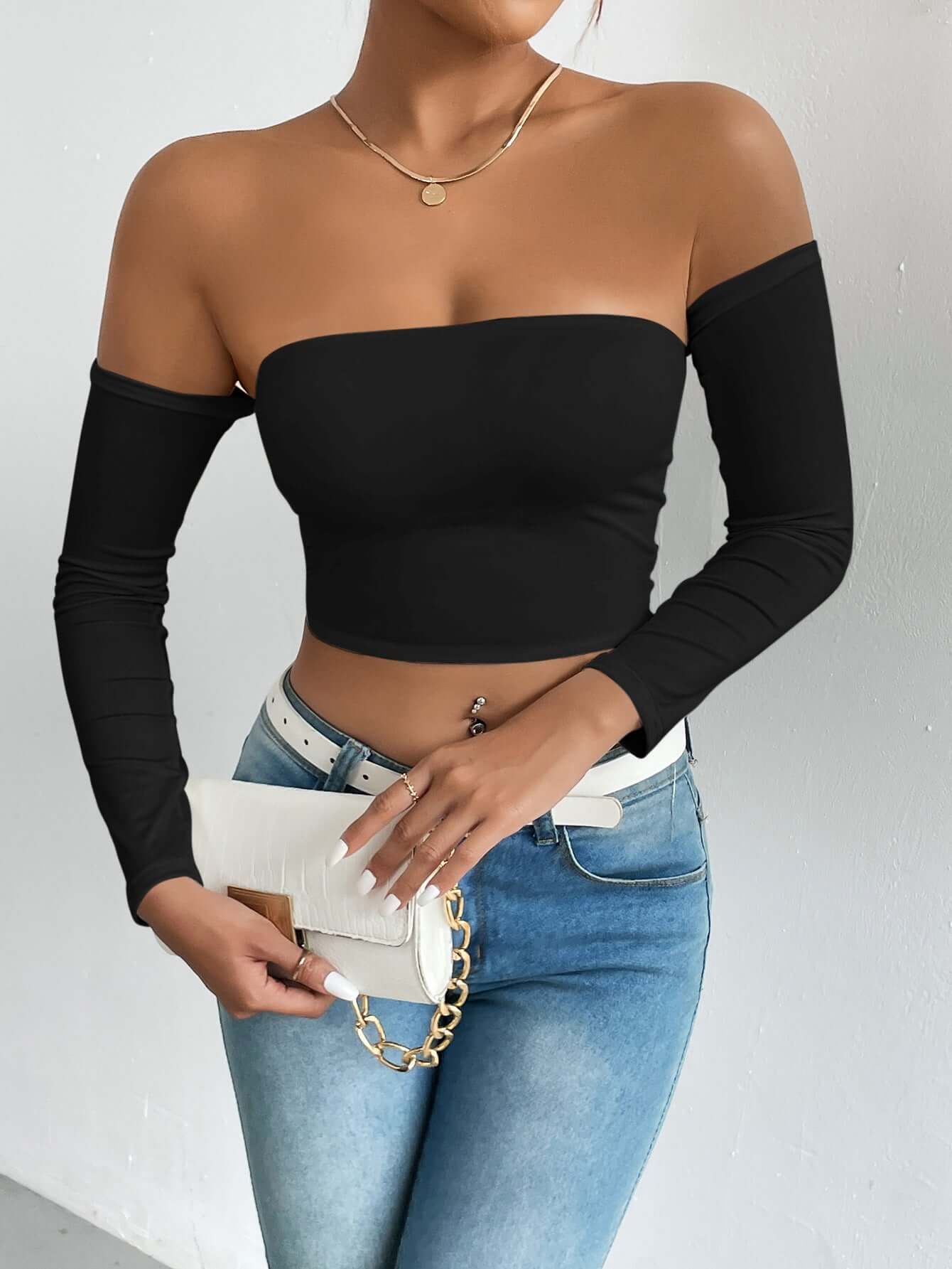 Woman wearing off-shoulder black top with blue jeans, holding a white clutch. Fashionable style showcasing casual elegance.