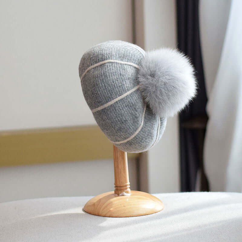 Stylish gray beanie with fur ball displayed on a wooden stand, perfect for keeping warm in winter.