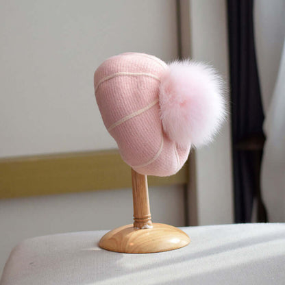 Pink beanie with a fluffy fur ball displayed on a wooden stand, perfect for winter style and warmth.