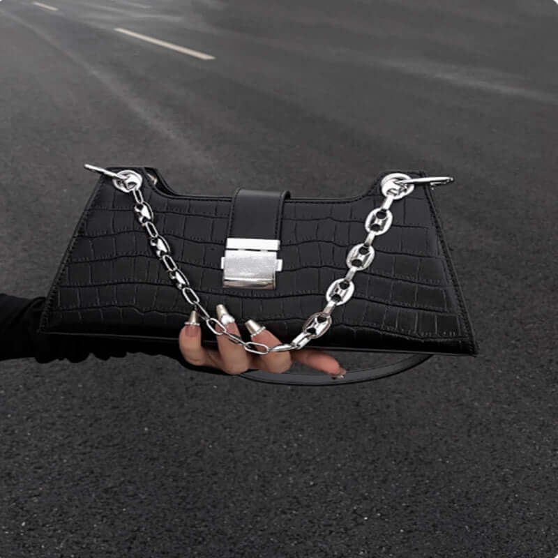 Black faux leather Baguette Handbag with stylish chain detail, held on street, highlighting its chic and slightly hard structure.