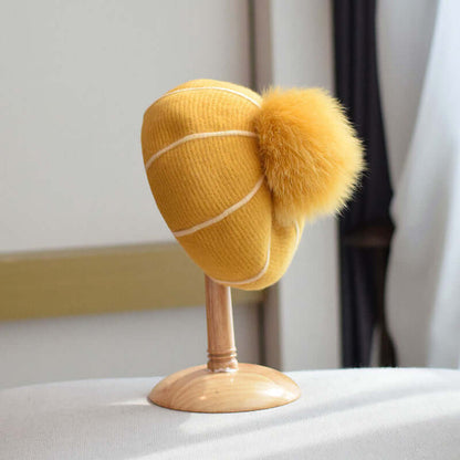 Cozy yellow beanie with fur ball displayed on a wooden stand, perfect for stylish winter wear.