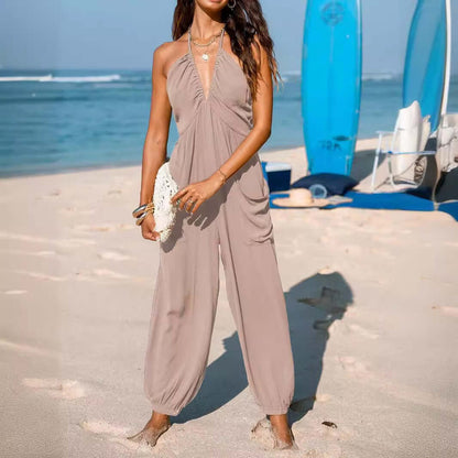 Woman wearing a back open jumpsuit in khaki, standing on a beach with surfboards and ocean in the background.