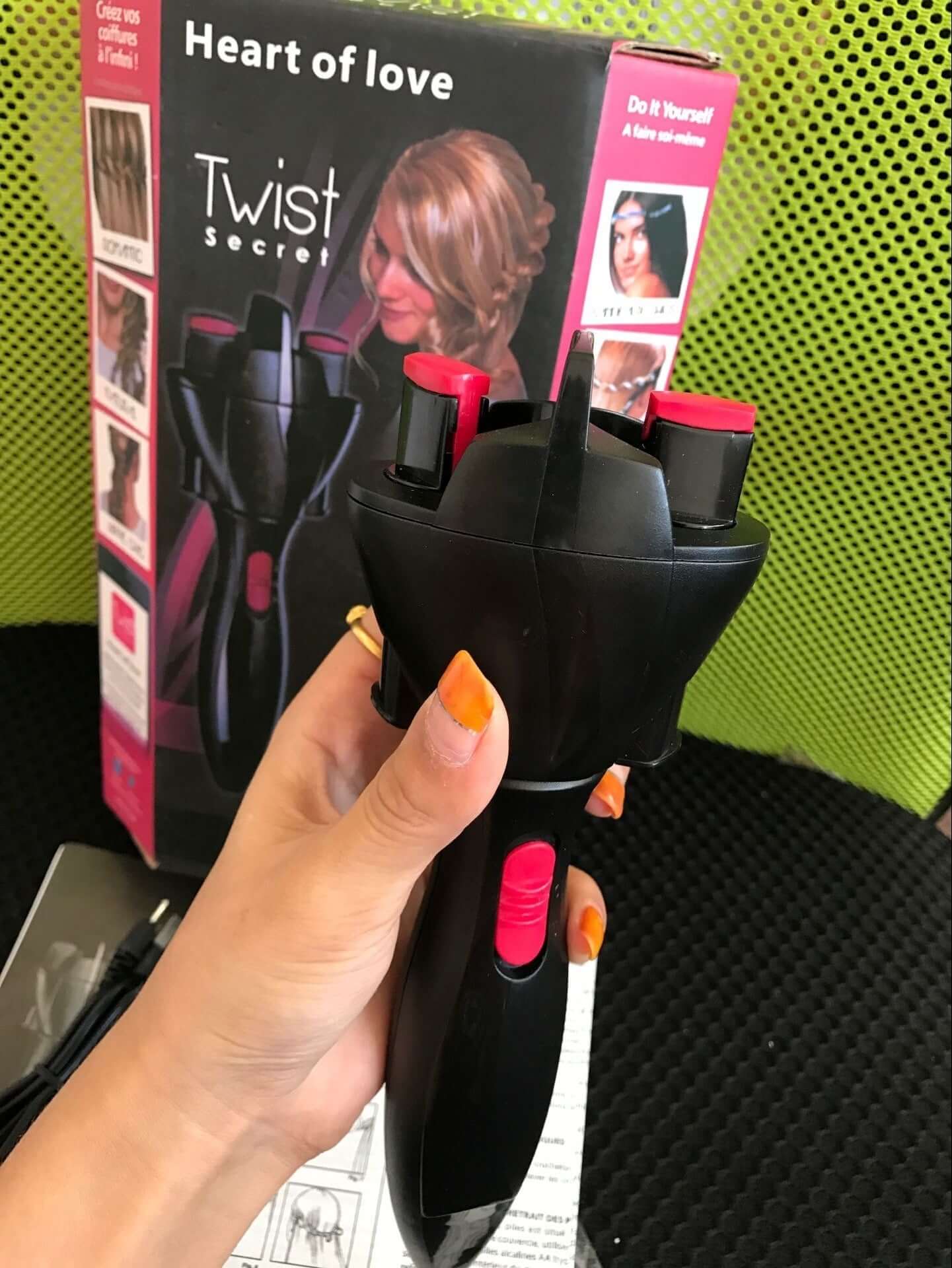 Person holding a compact machine for braiding hair with pink accents, shown with packaging box in the background.