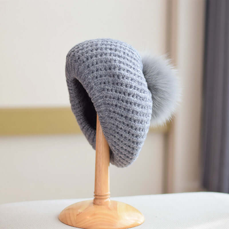 Elegant grey Fur Ball Hat displayed on a wooden stand, featuring a cozy design and stylish pom-pom for winter wear.